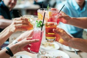 Ao centro aparecem seis copos com bebidas em um momento de brinde